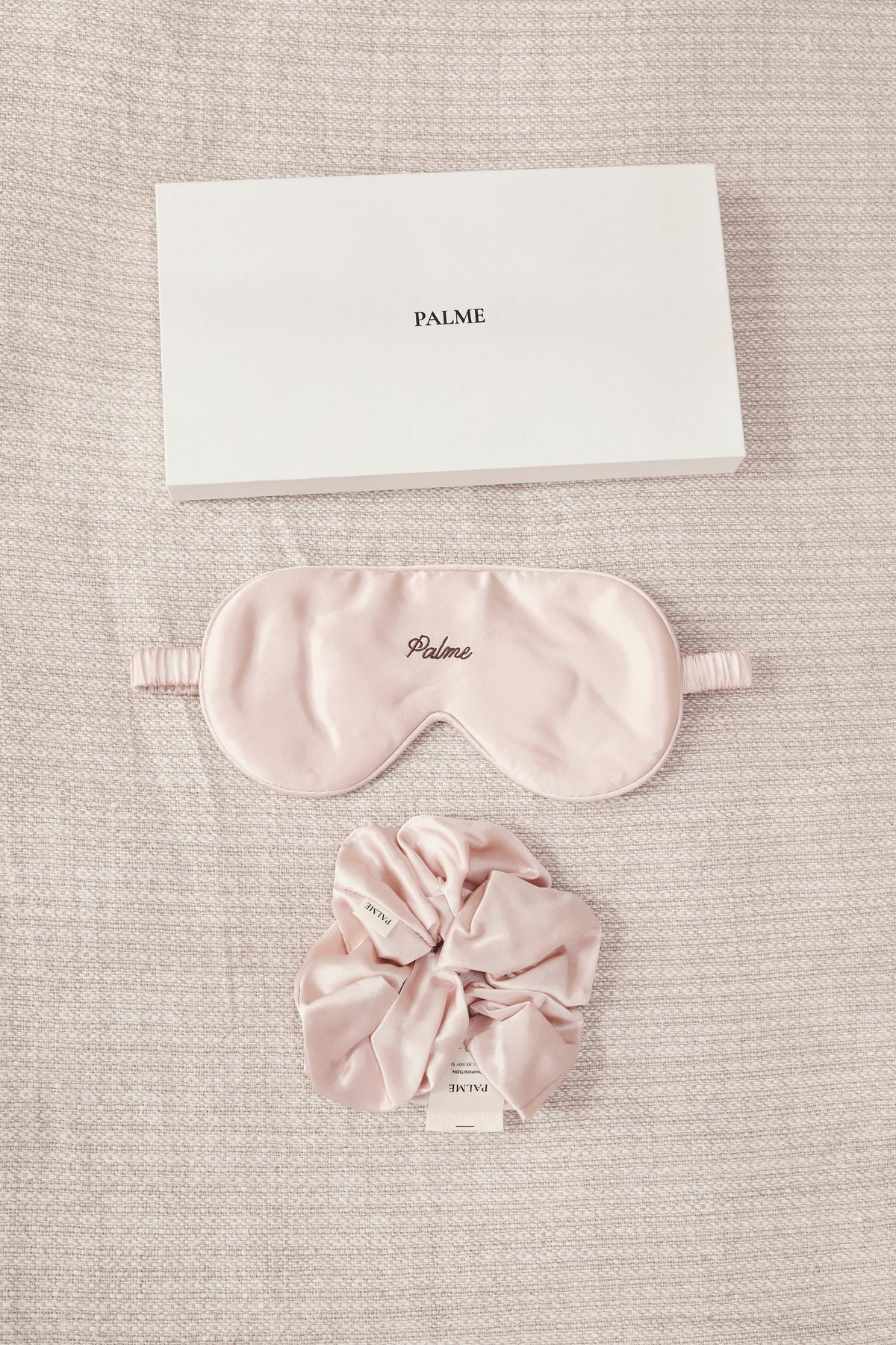 A dusty pink silk sleep mask and matching scrunchie set with a white rectangular PALME box, arranged on a textured beige linen background.