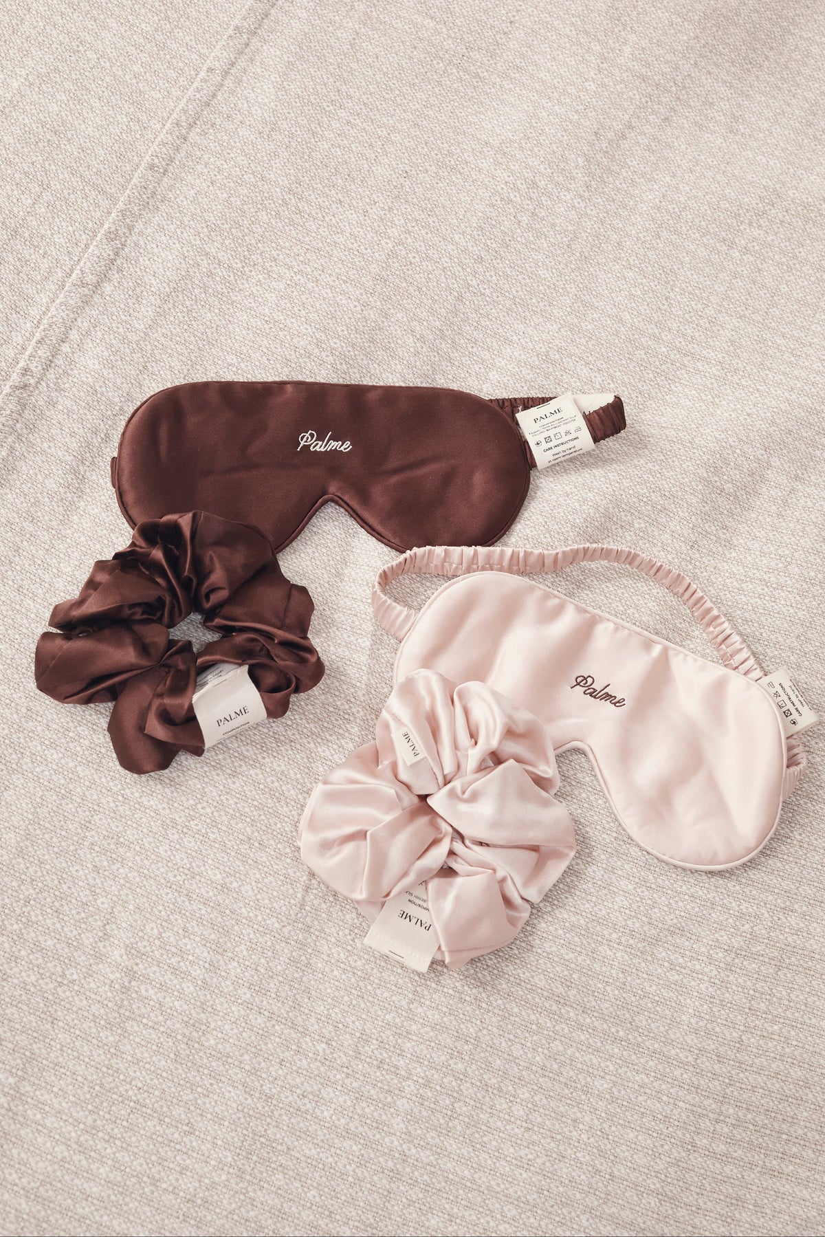 Two satin sleep masks and two matching scrunchies in brown and blush pink, laid flat on a textured beige fabric background. Each set features a mask and scrunchie with the brand name 'PALME' embroidered on it.
