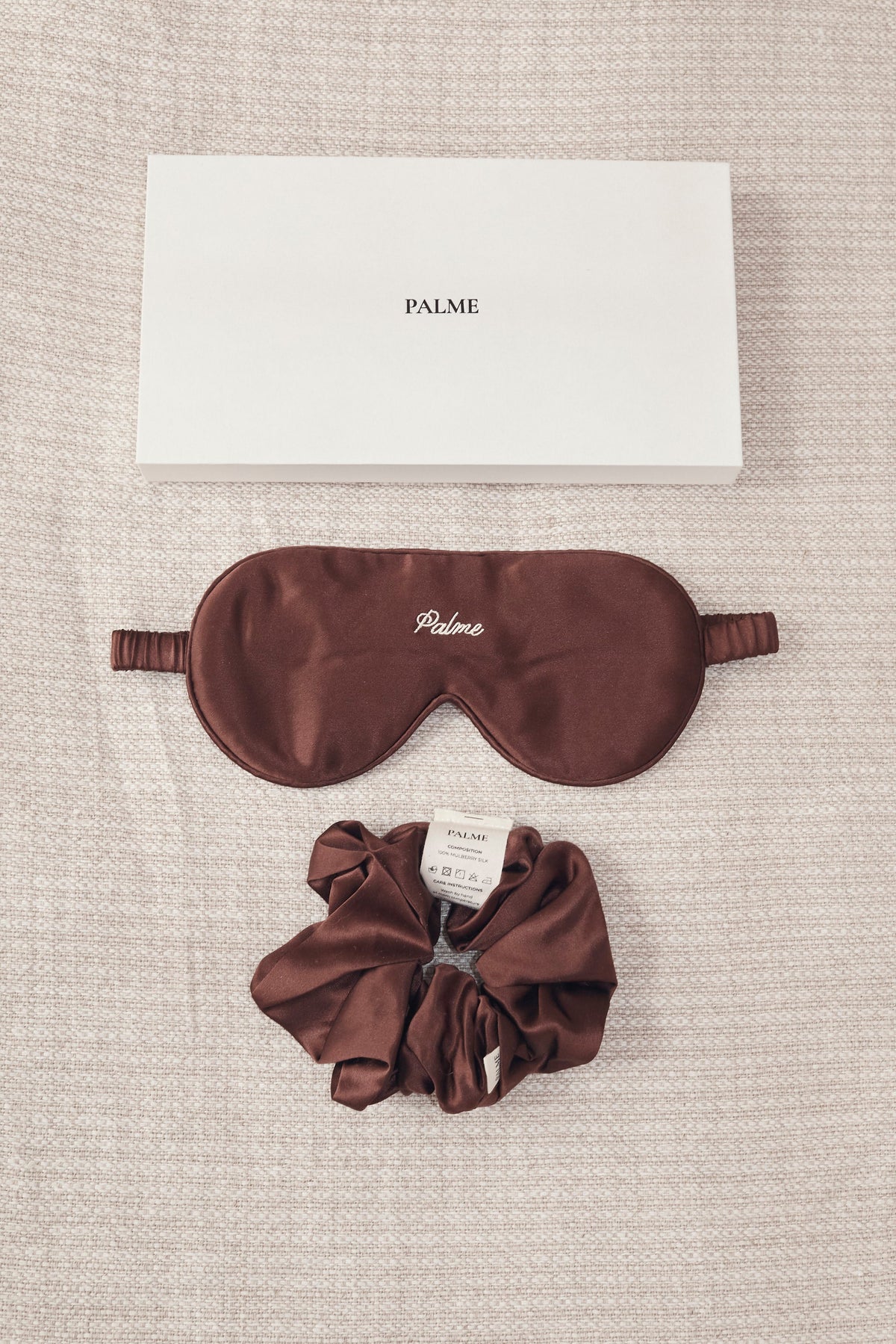 A brown silk sleep mask and matching scrunchie set with a white PALME brand box, arranged on a textured beige linen background.