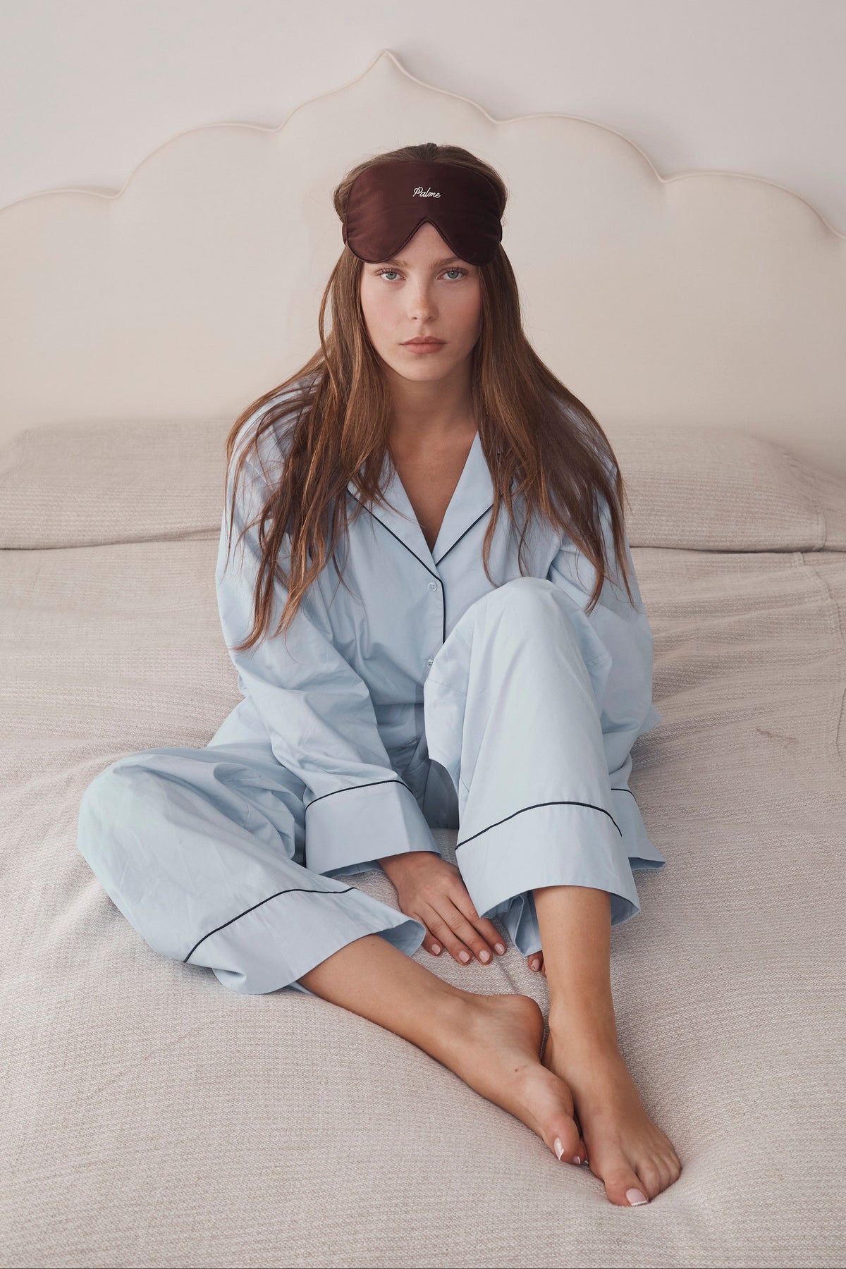 A woman sits on a bed wearing a light blue pajama set with dark piping and a dark brown sleep mask. The background features a cream-colored headboard and neutral bedding.